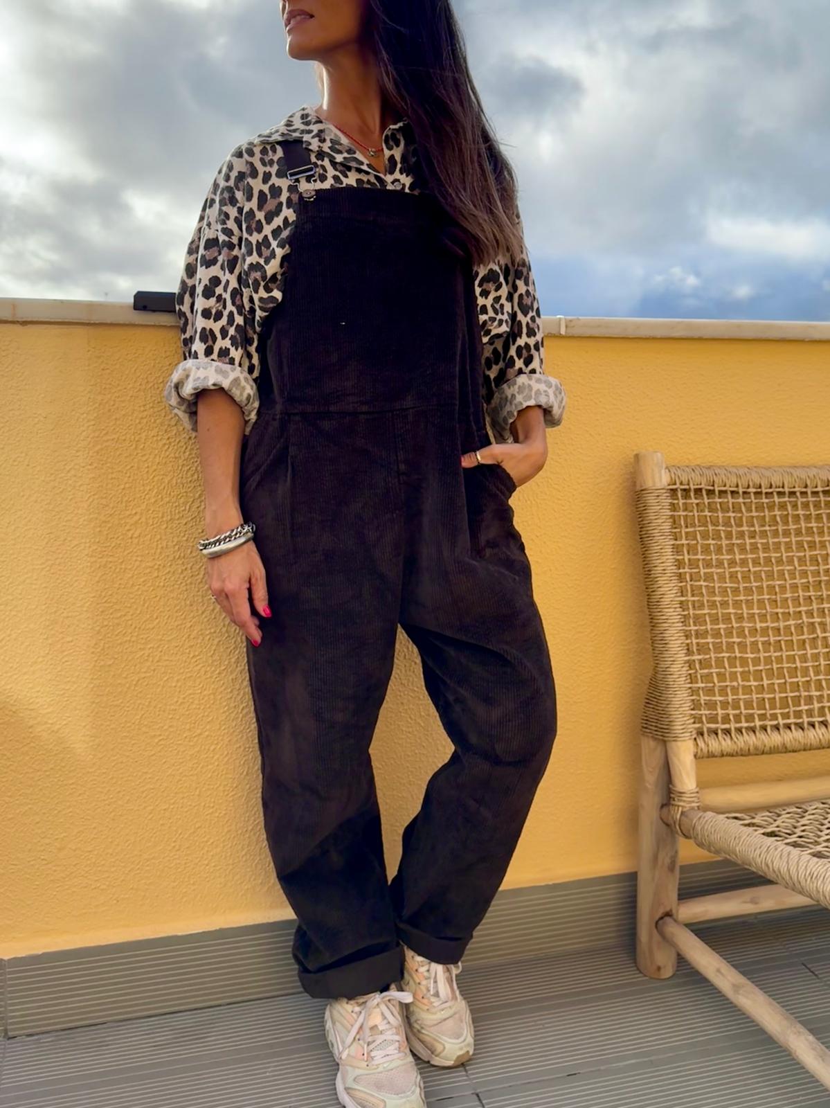 Mulher com macacão preto e camisa estampada animal print junto a parede amarela e cadeira de madeira com corda entrelaçada