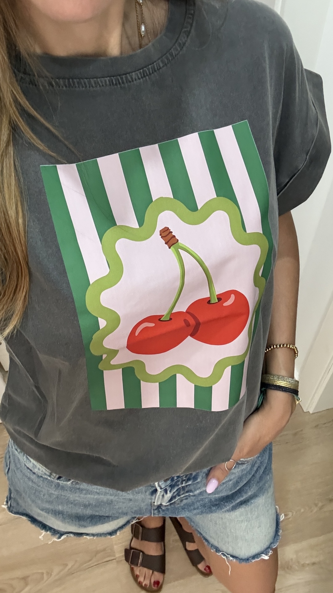 T-shirt cinza escuro com estampado de cerejas e riscas verdes e brancas, calção de ganga azul claro e sandálias castanhas