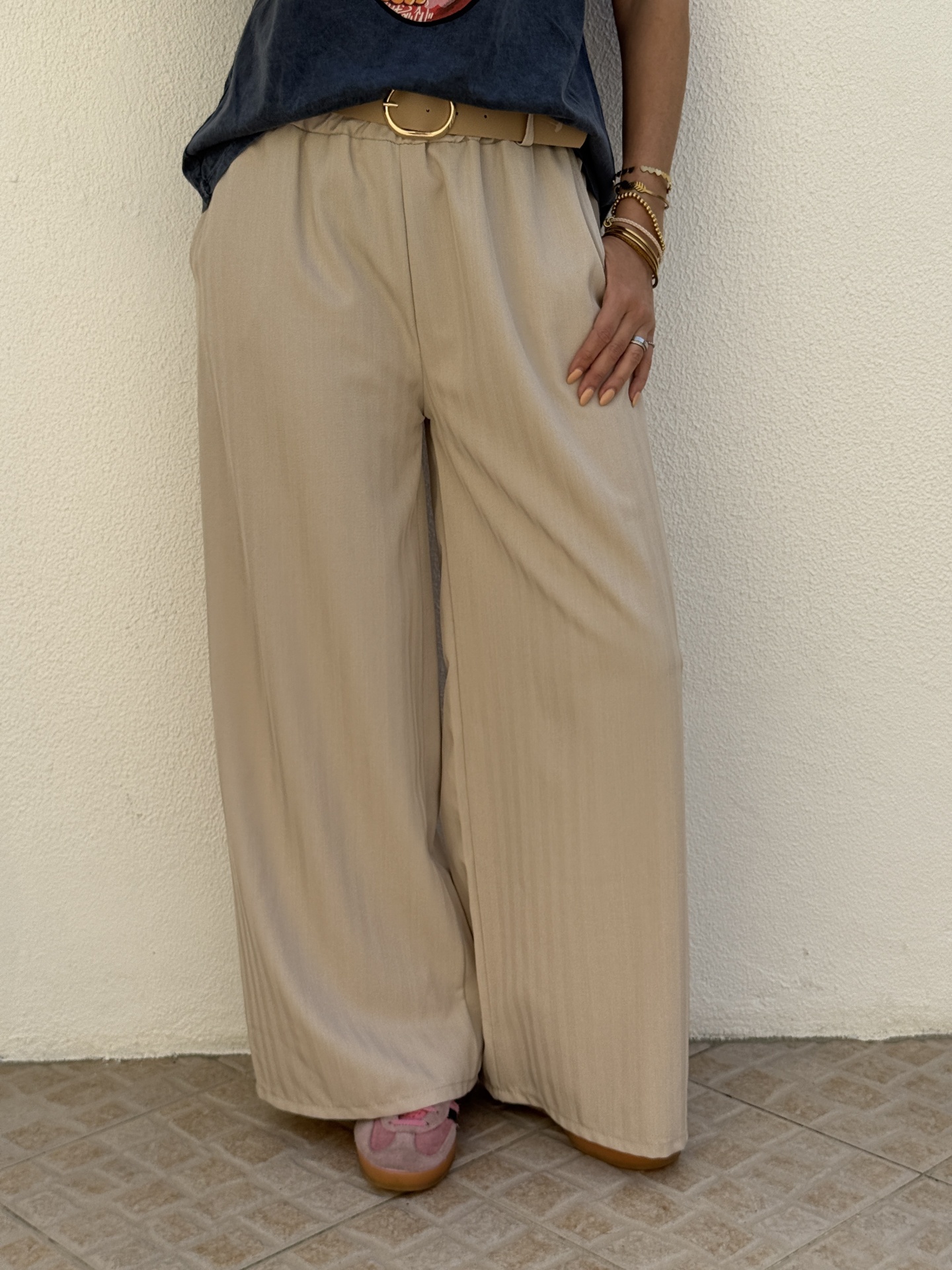 Calças largas bege com cinto castanho claro, parte de cima azul escuro, acessórios dourados, parede branca e chão de azulejos.