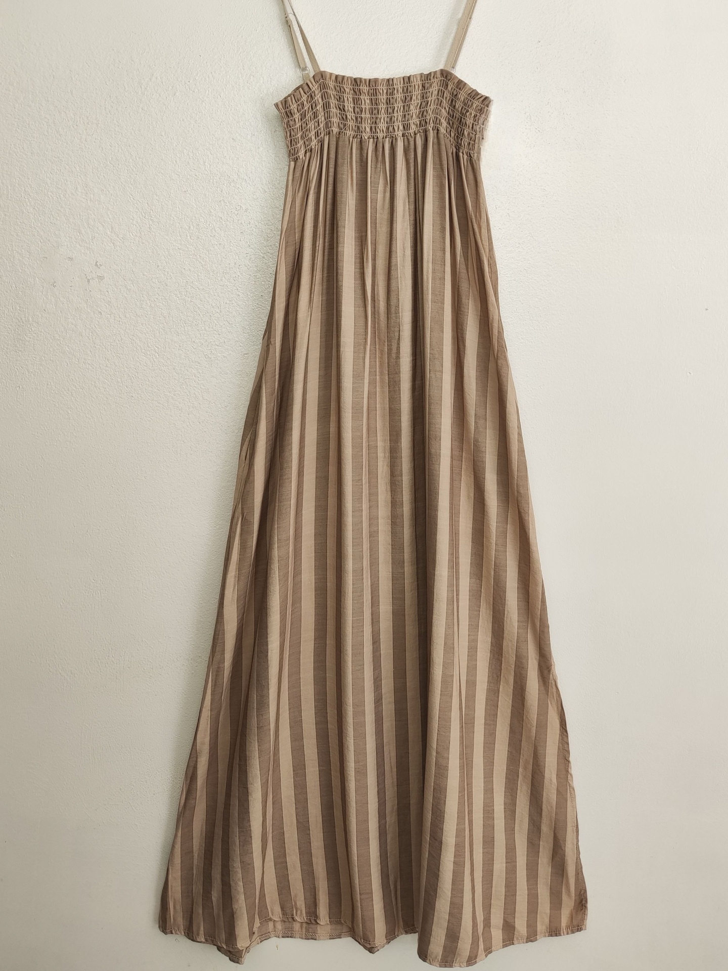 Vestido castanho claro plissado com alças finas pendurado numa parede branca