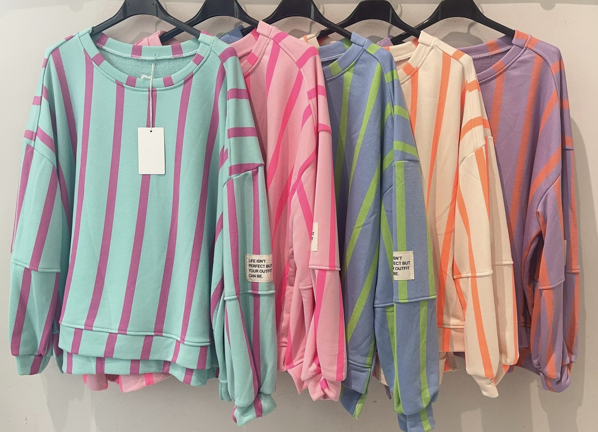 Cinco camisolas às riscas coloridas penduradas em cabides pretos com etiqueta branca com texto em preto