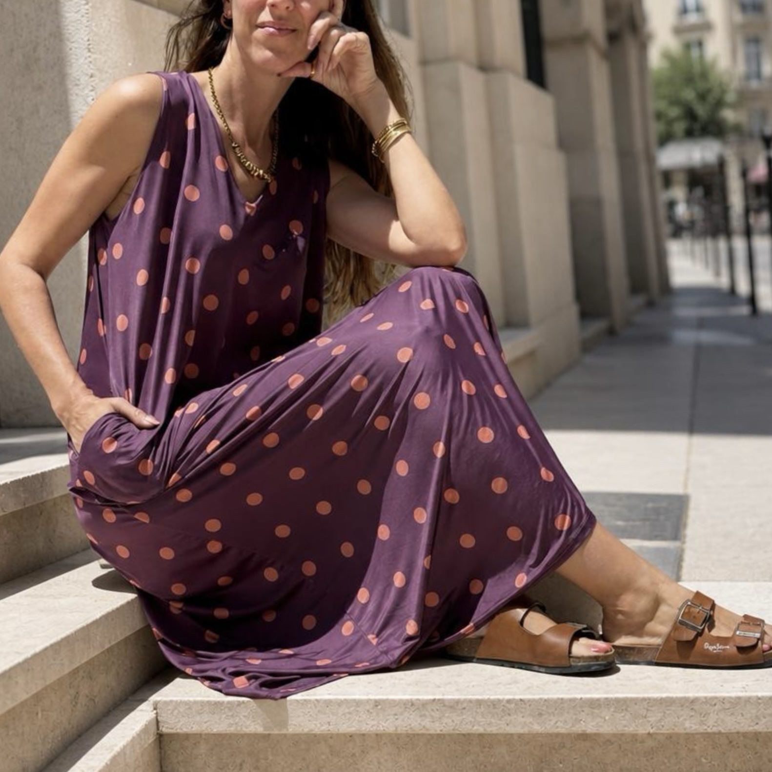 Mulher com vestido roxo de bolinhas sentada em degraus com sandálias castanhas