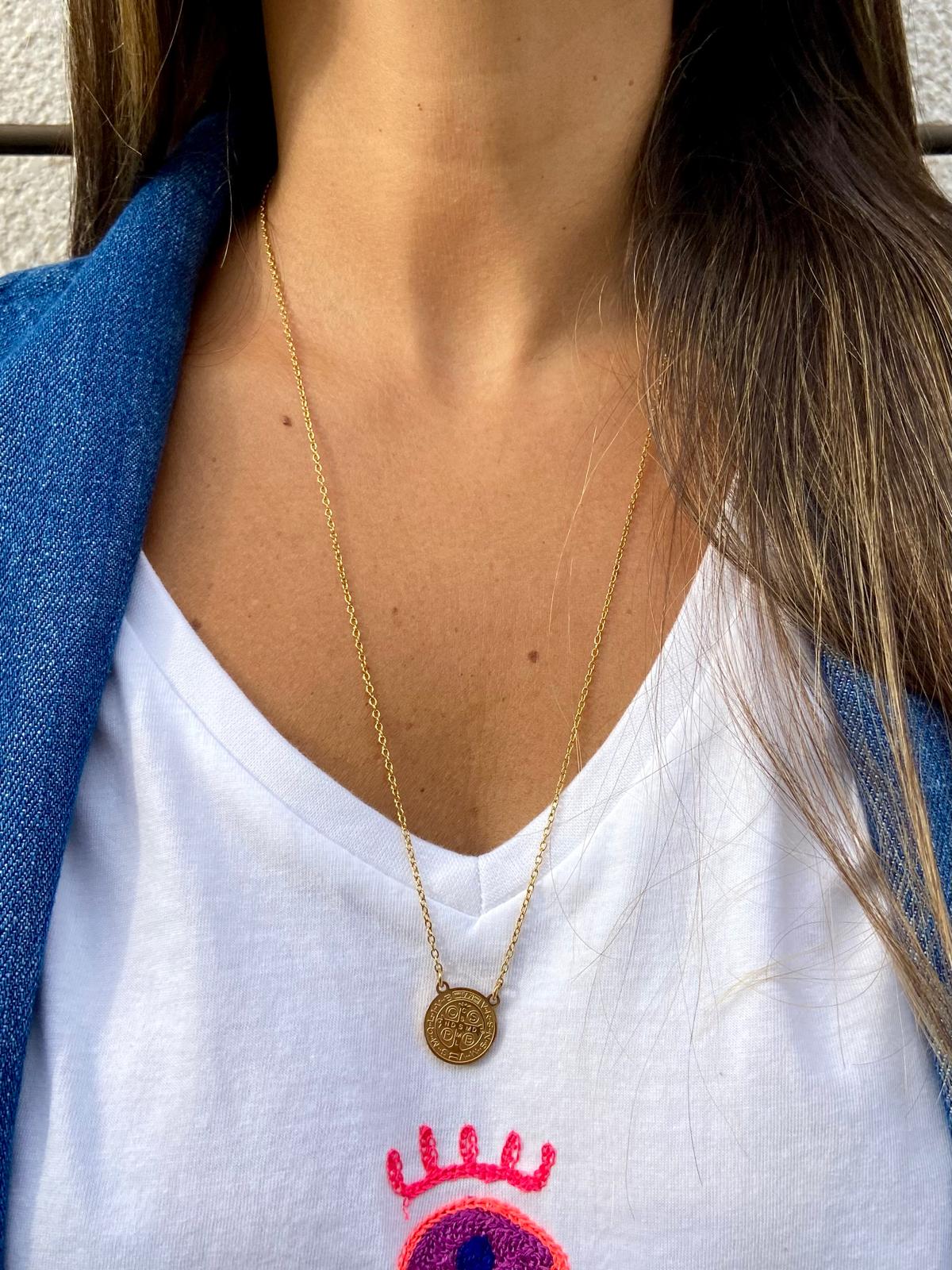 Colar dourado com pendente redondo numa pessoa com t-shirt branca com estampa de olho e coroa e casaco azul