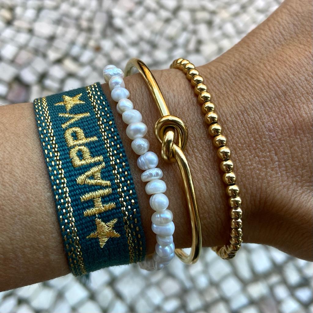 Quatro pulseiras no pulso, tecido verde com dourado, pérolas brancas, dourada com nó, contas douradas
