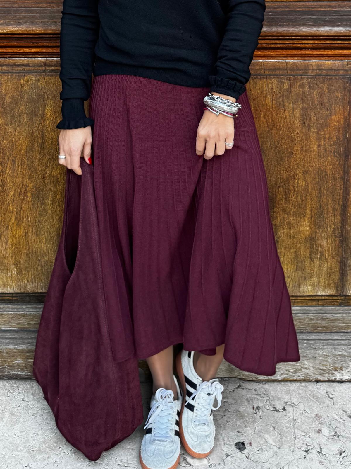 Saia midi plissada vinho com ténis brancos e blusa preta, pulseiras e fundo de madeira.