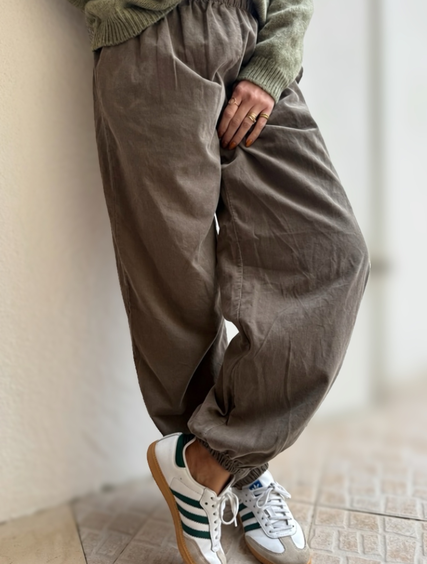 Pessoa vestindo calças cinza largas e ténis brancos com riscas verdes perto de parede clara