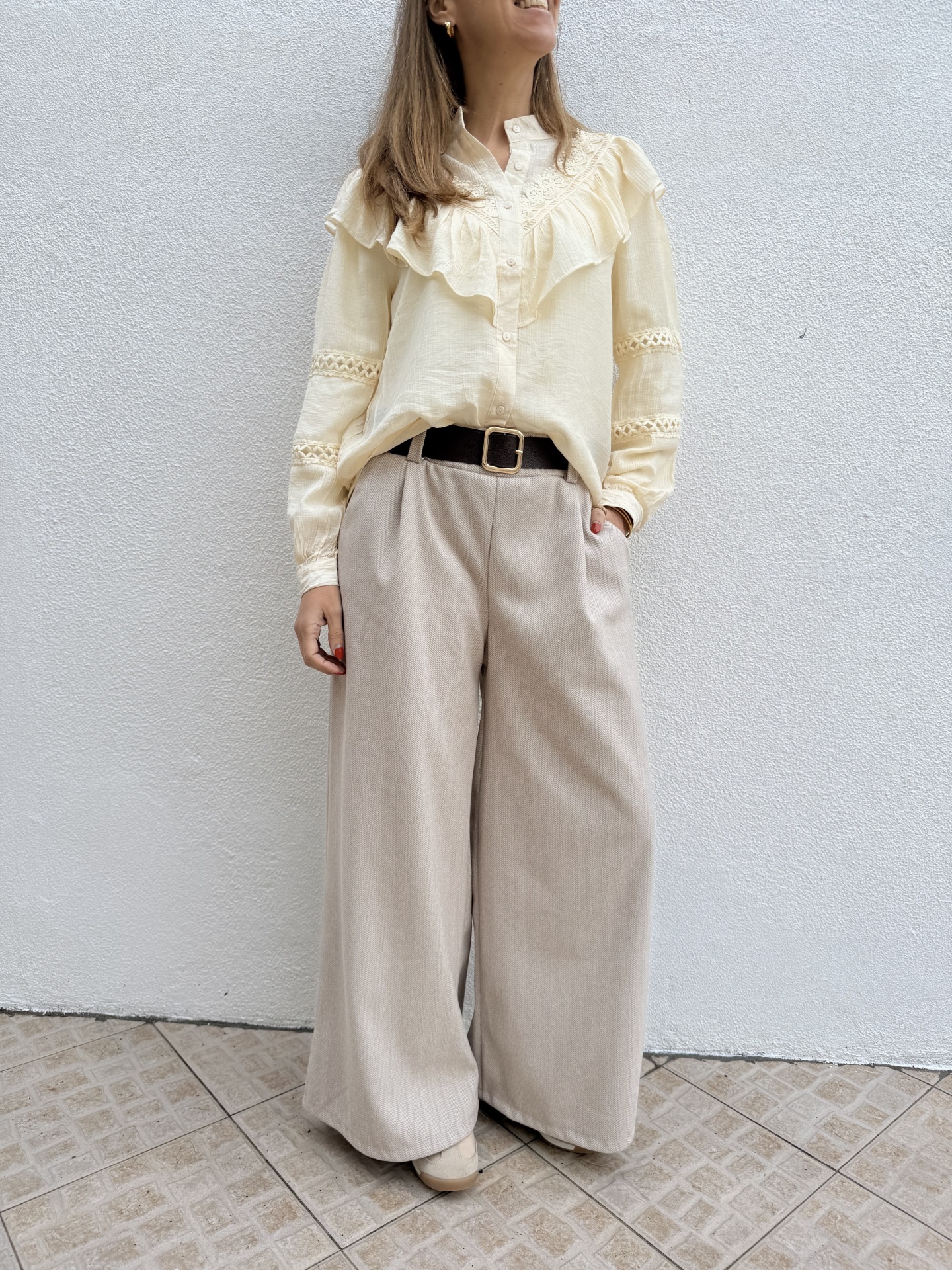 Blusa creme com folhos e bordados e calças largas bege claras com cinto preto.