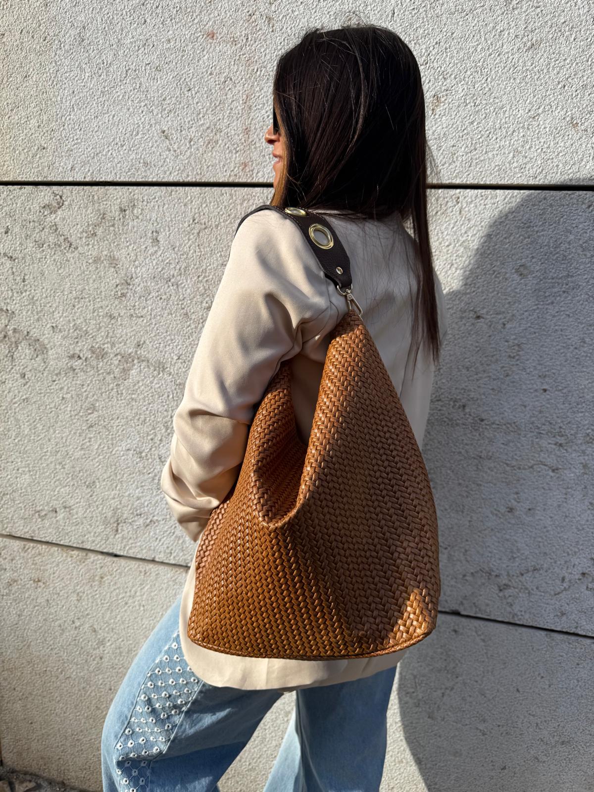 Mala de pele castanha com padrão tecido e alça preta metálica usada por mulher junto a parede clara
