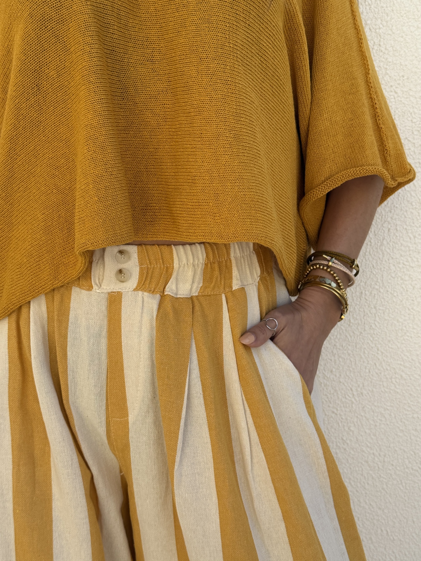 Topo amarelo mostarda de malha e calças riscas verticais amarelo e cru com botões na cintura, pulseiras no pulso.