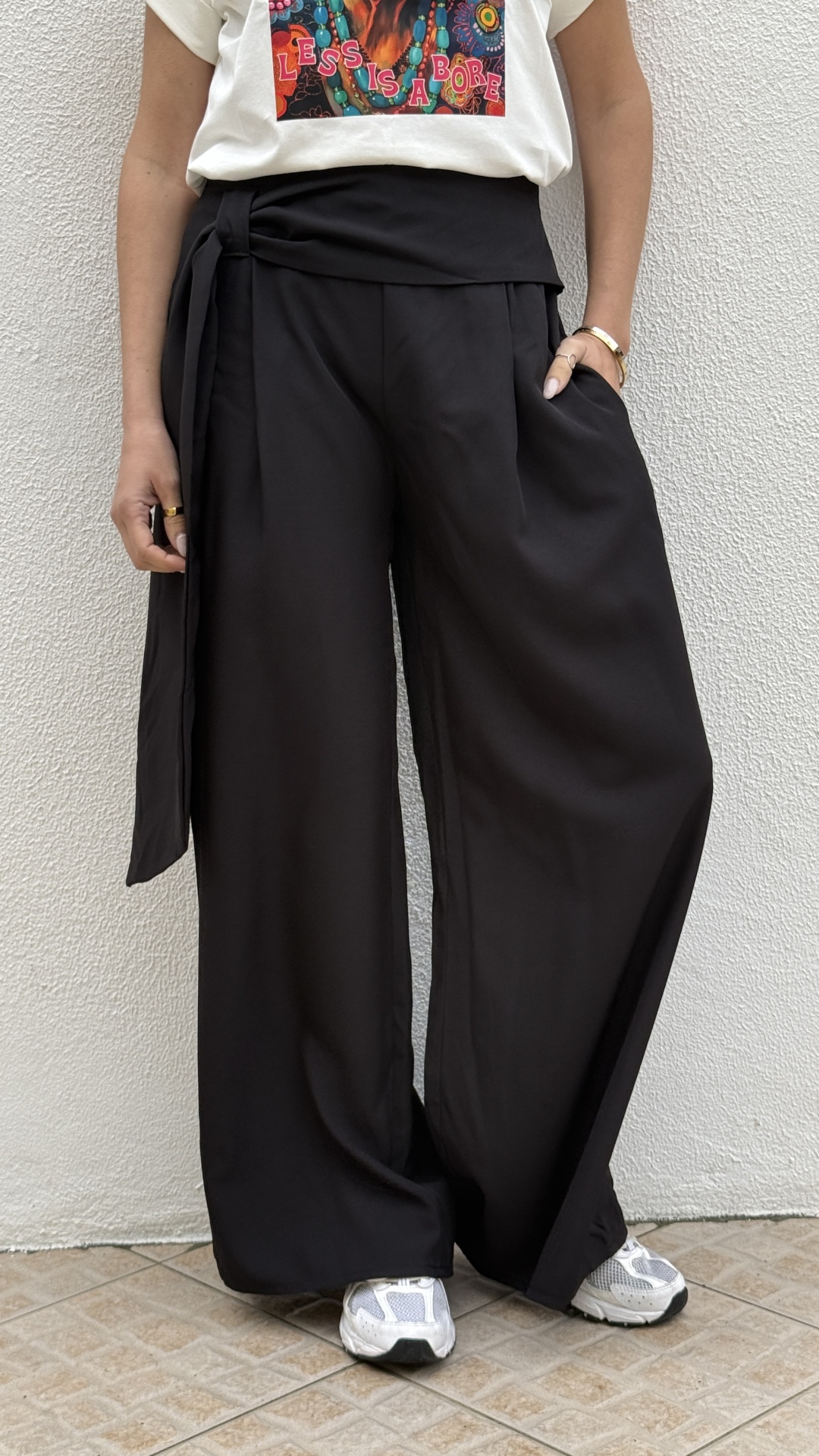 Calças pretas largas com cós largo e faixa, t-shirt branca com estampa com texto, ténis brancos junto a parede branca