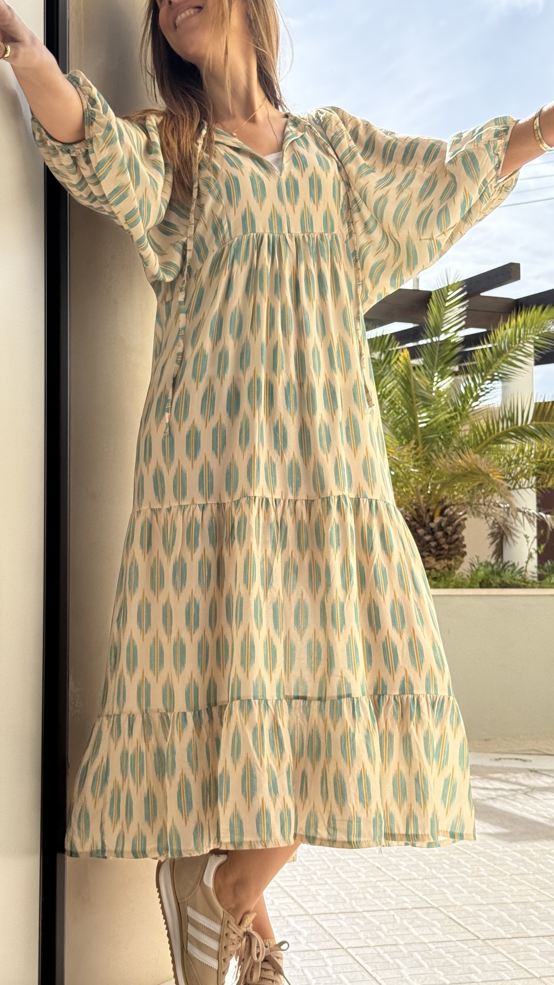Vestido comprido com padrão geométrico bege e verde esbatido junto a parede e ténis bege.