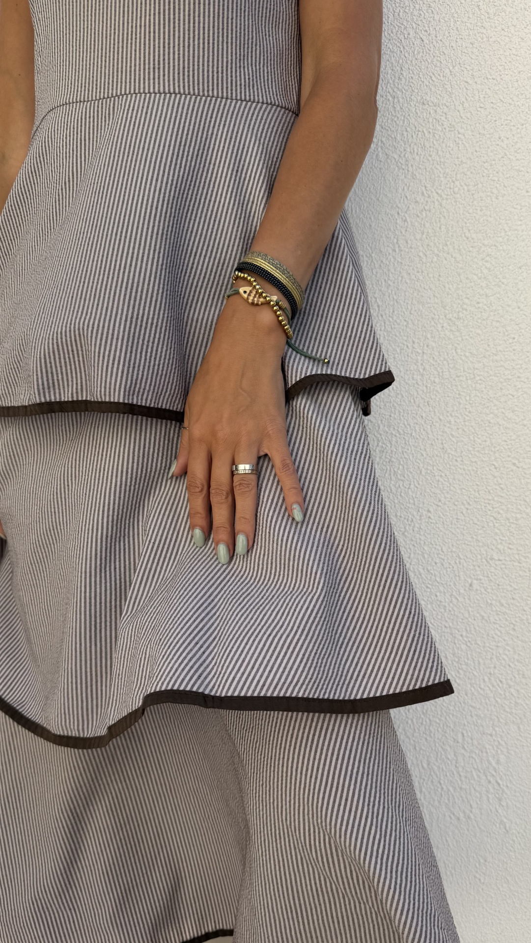 Vestido risca de giz cinzento e branco com acabamento castanho, pulseiras e anel, mão feminina.