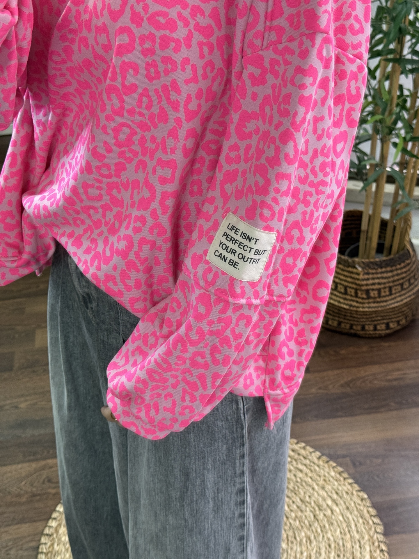 Sweatshirt rosa com padrão de manchas rosa e etiqueta com texto na manga, calças de ganga cinza e planta em vaso.