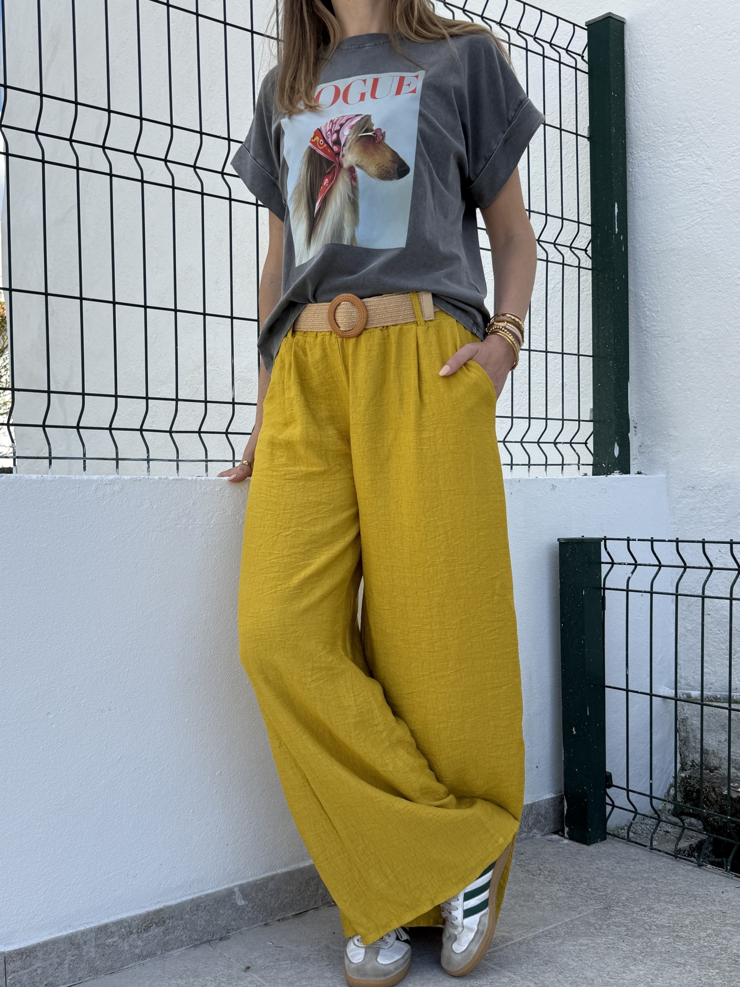 Pessoa vestindo camisola cinzenta com estampa de cão e calças largas amarelas, com cinto bege e sapatilhas brancas com riscas verdes.