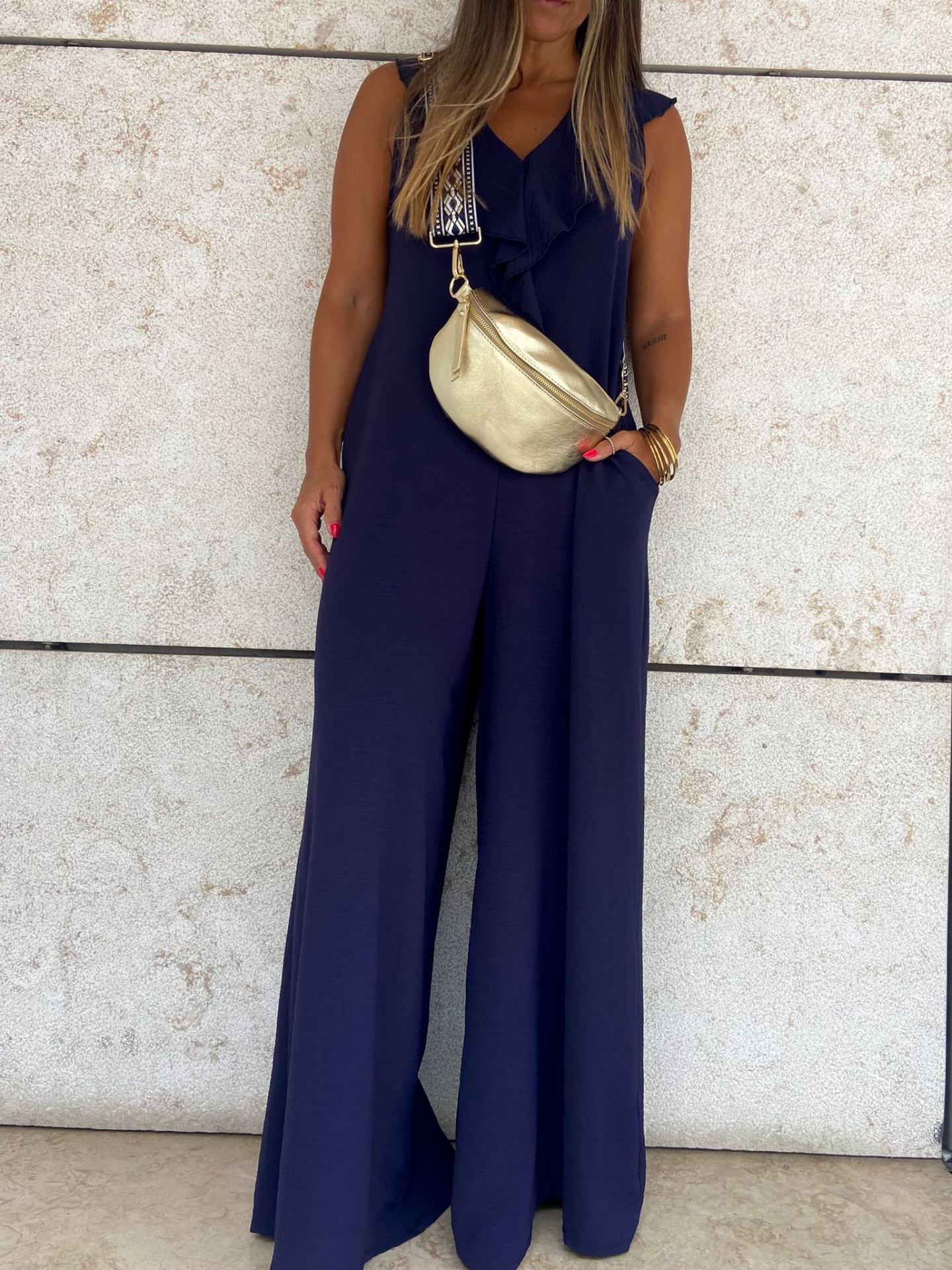 Mulher com macacão azul e bolsa tiracolo dourada em fundo de parede beige