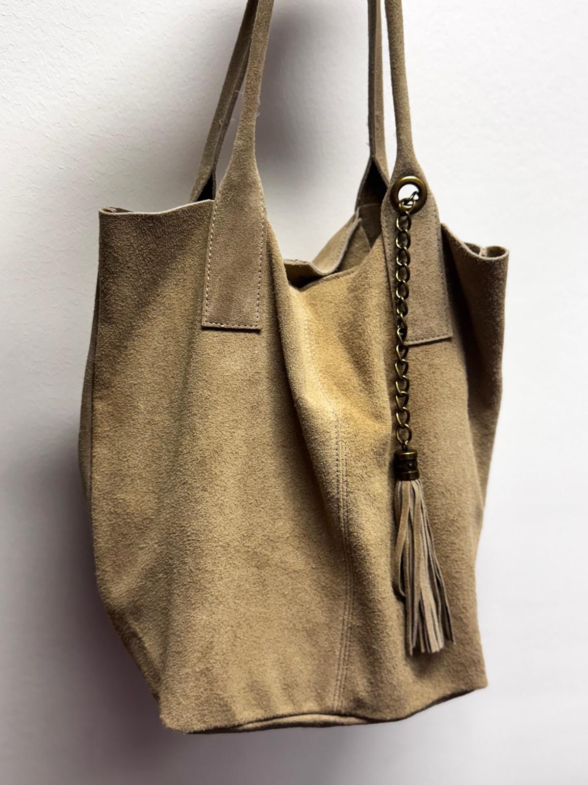 Mala de camurça bege com alças duplas e pendente tassel em corrente, pendurada numa parede branca