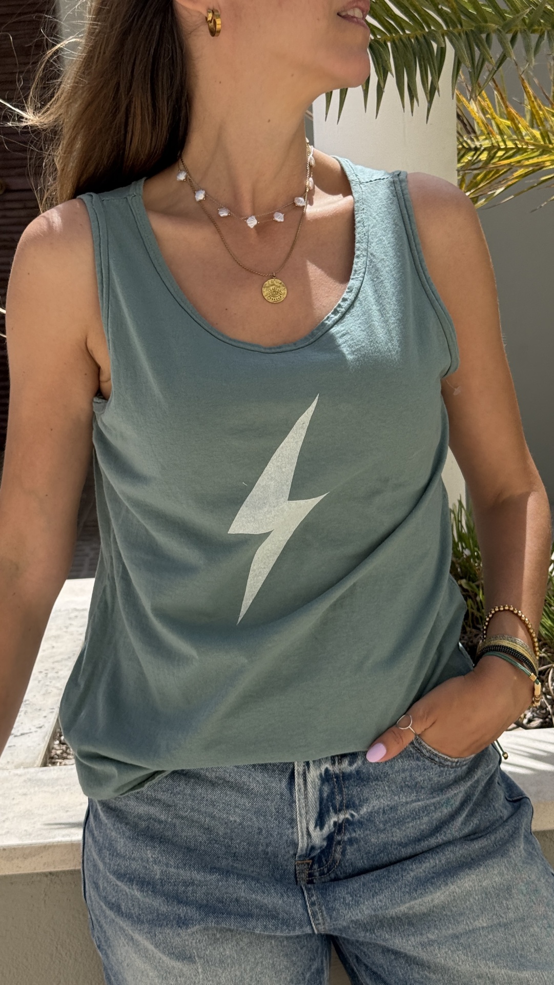 Mulher com t-shirt verde-azulada com símbolo de raio, jeans e acessórios dourados em ambiente com plantas