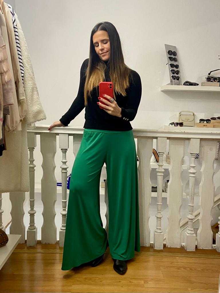 Mulher com camisola preta e calças verdes largas posando em ambiente interior com corrimão branco e óculos de sol.