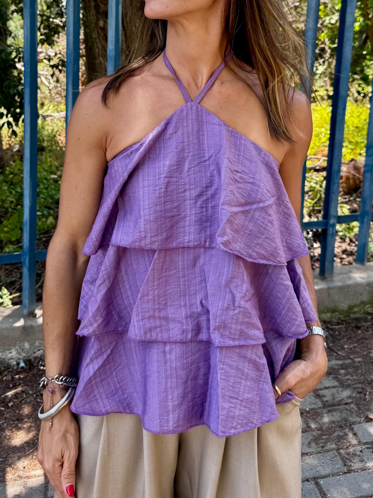 Top feminino lilás com babados e alças finas, calças bege, ambiente exterior com vegetação e grade azul