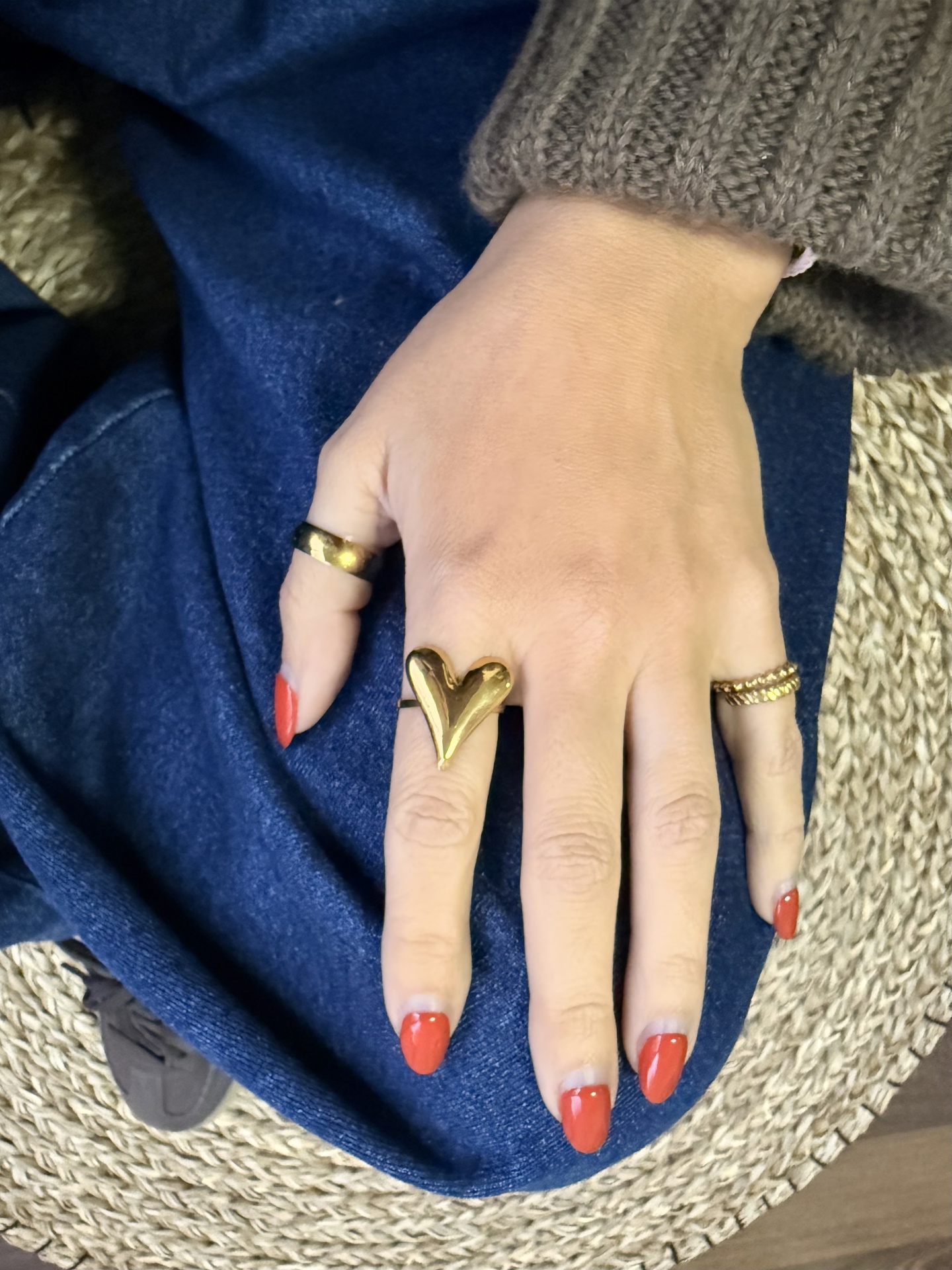 Mão feminina com três anéis dourados e unhas vermelhas sobre tecido azul e ráfia