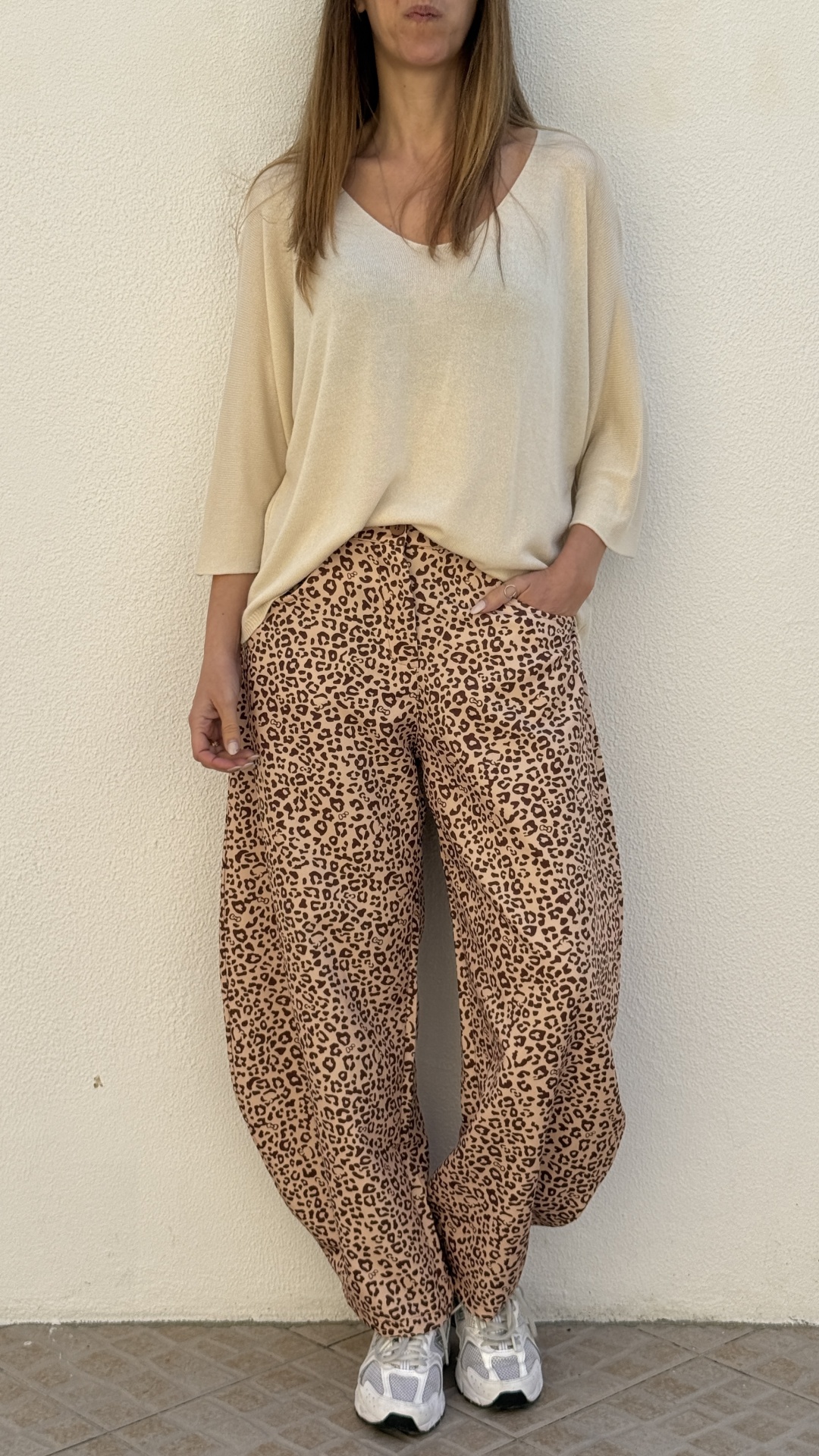 Pessoa com calças de estampa animal print, camisola bege clara e sapatilhas brancas e cinza de pé junto a uma parede branca.