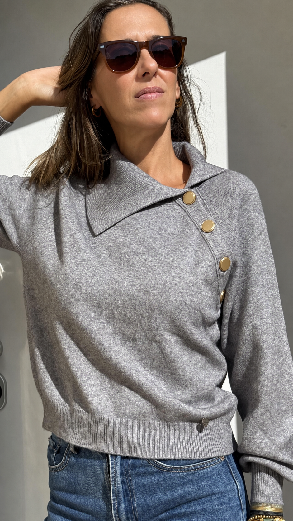 Mulher com camisola cinza de gola larga com botões dourados e jeans azul a usar óculos de sol castanhos