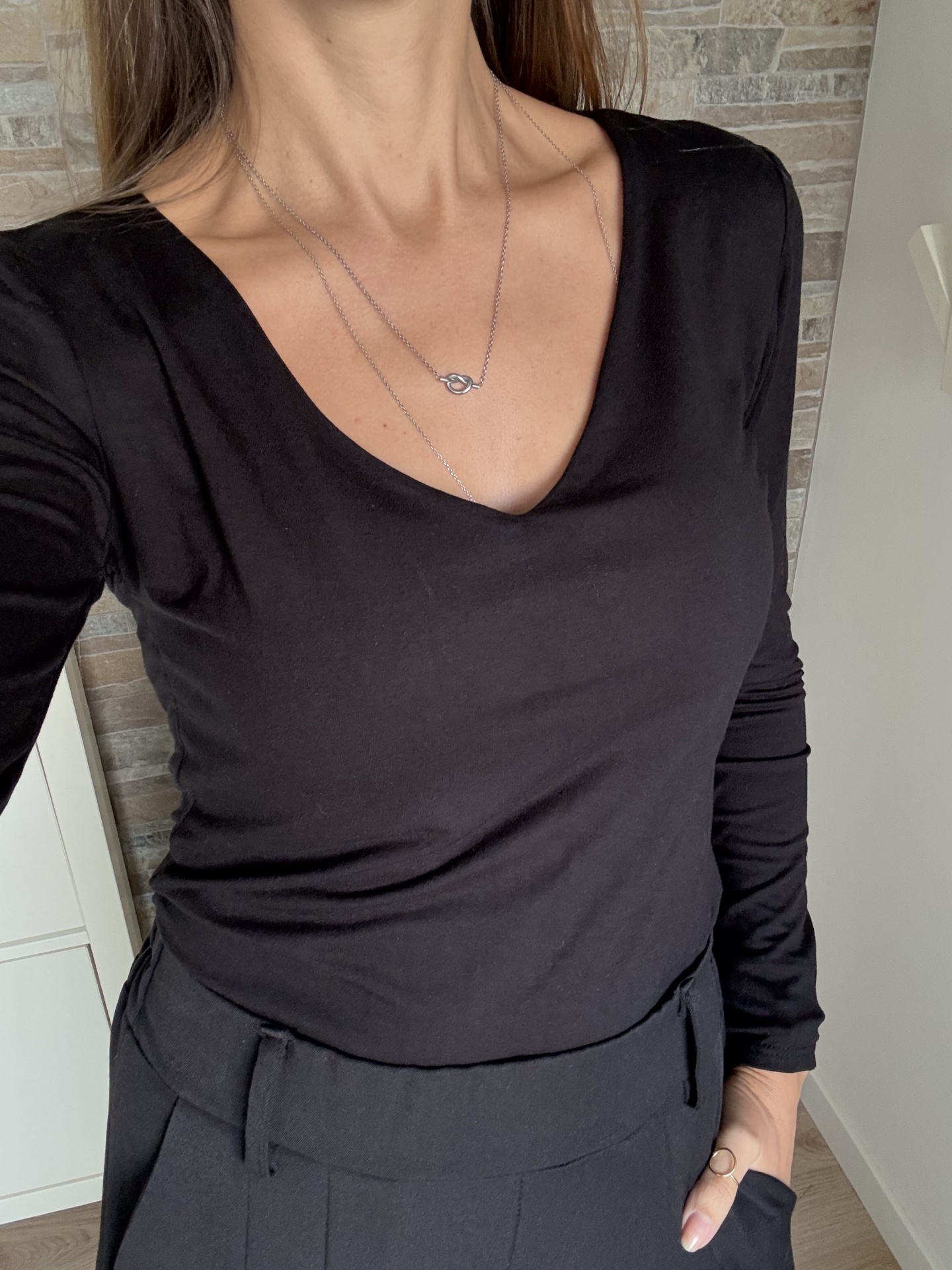 Mulher vestindo camisa preta de manga longa com decote em V, calças pretas e colar prateado com pendente de nó