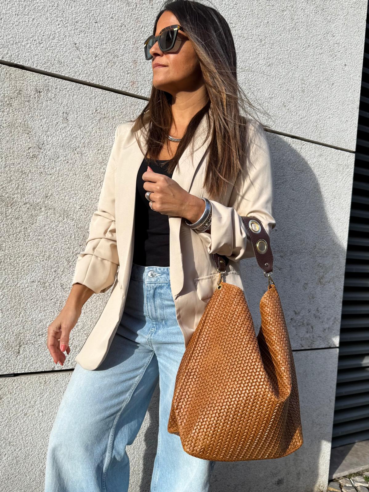 Mulher com casaco bege, top preto, calças de ganga azuis e mala castanha entrelaçada ao ar livre