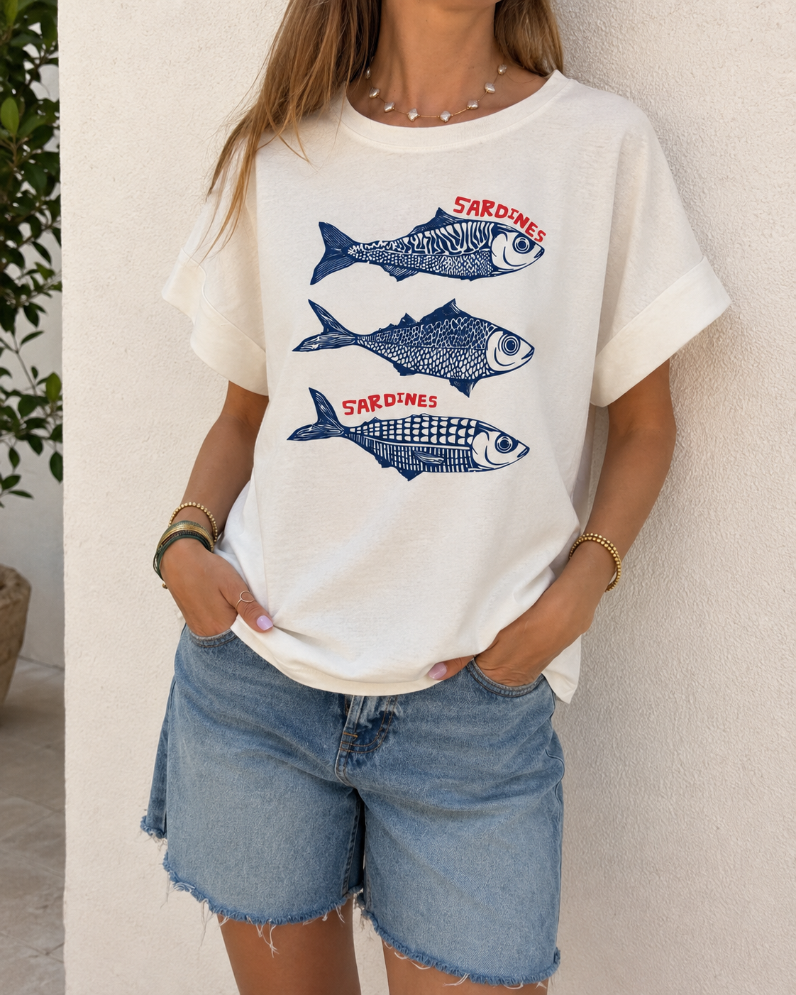 T-shirt branca com estampado de sardinhas azuis e texto vermelho, calções de ganga azuis, cenário exterior com parede branca e planta