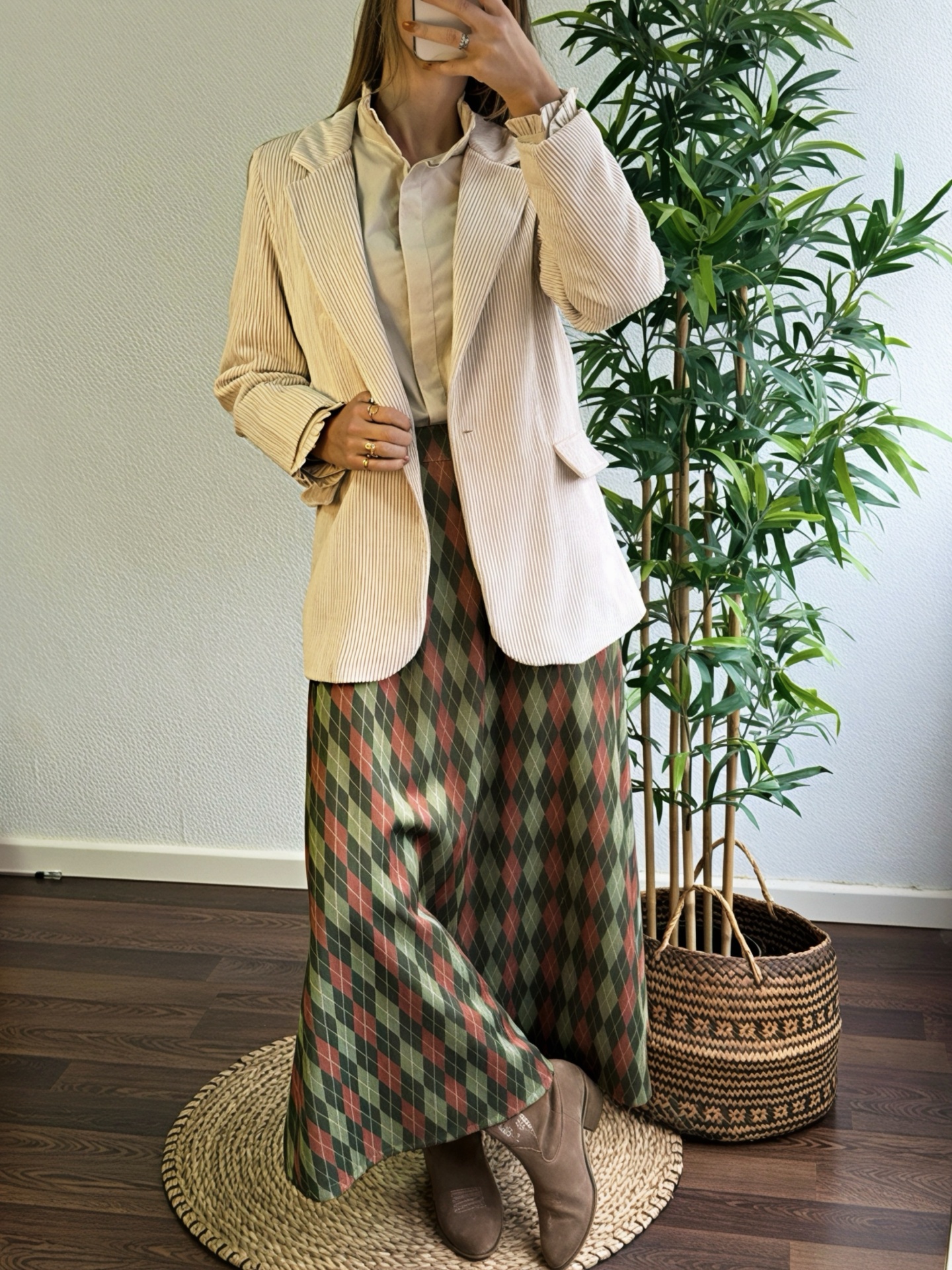 Pessoa a usar blazer bege de veludo cotelê, camisa bege, saia xadrez verde e castanha, botas castanhas, ao lado de planta dentro de cesto de palha