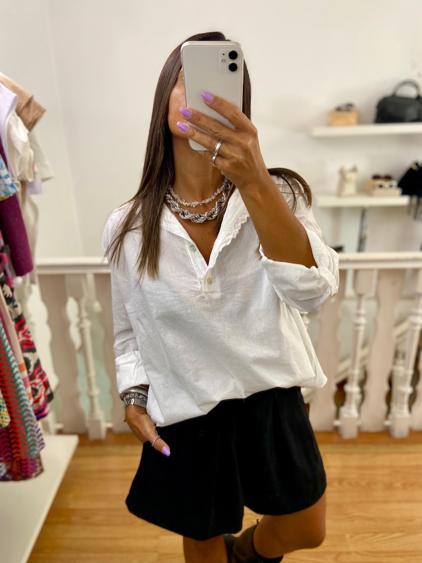 Pessoa a tirar selfie com telemóvel branco com camisa branca e calções pretos em interior com chão de madeira.