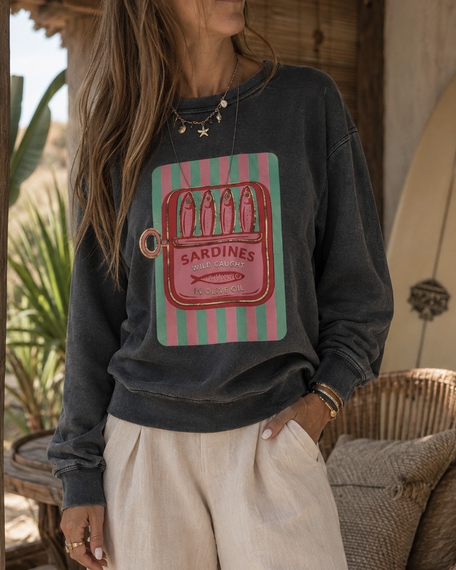 Camisola cinza escura com estampa colorida de lata de sardinhas e calças claras num ambiente interior acolhedor