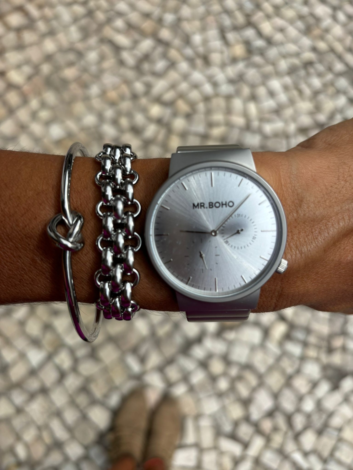 Relógio MR.BOHO prata com pulseiras metálicas num pulso em fundo de calçada portuguesa