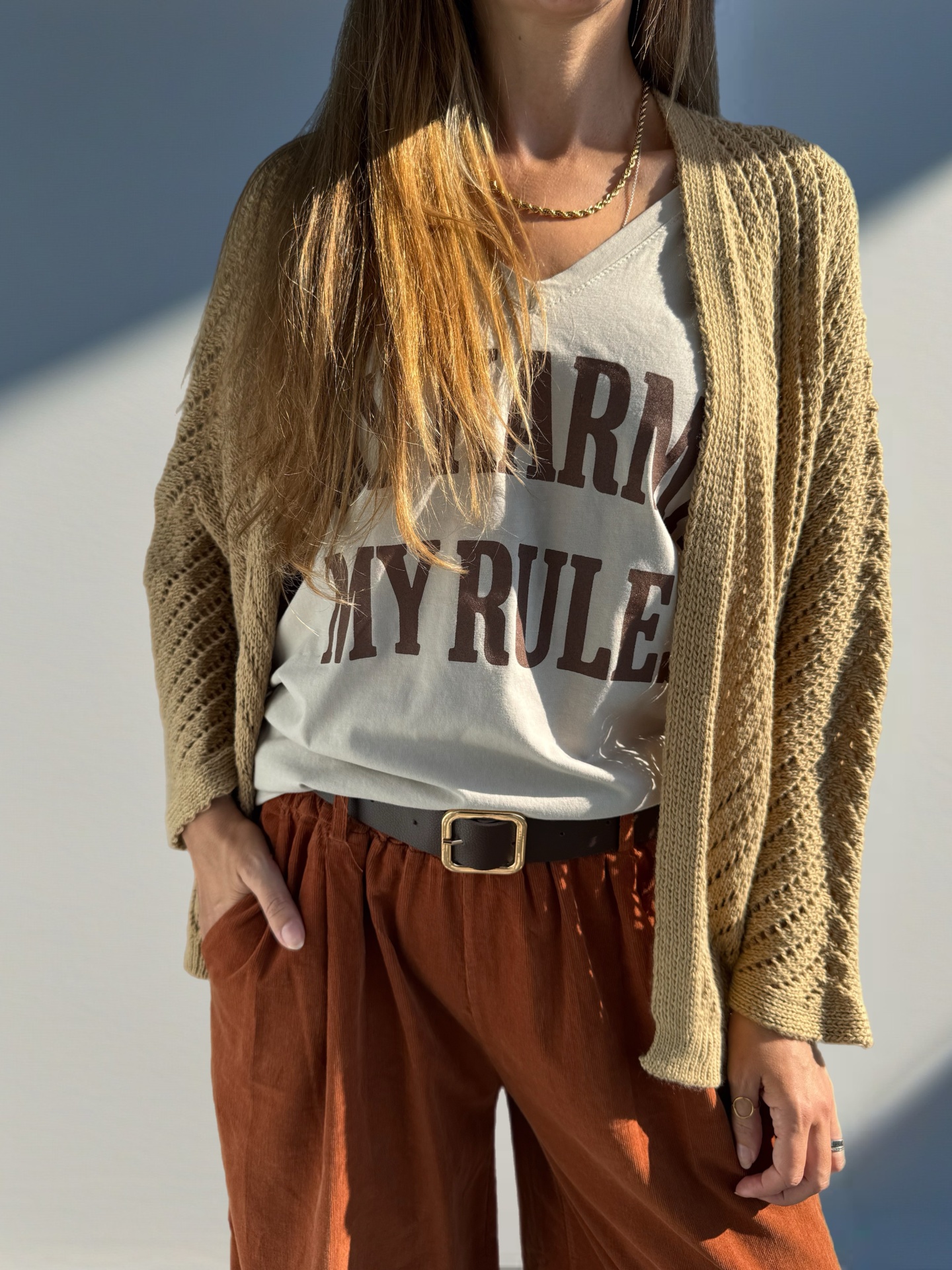Top branco com texto, cardigã bege, calças castanhas e acessórios