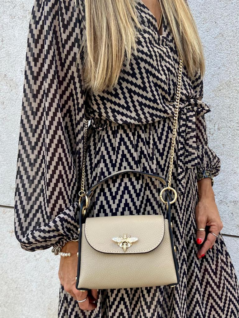 Bolsa feminina pequena bege com corrente dourada e emblema de abelha, usada com vestido padrão preto e bege