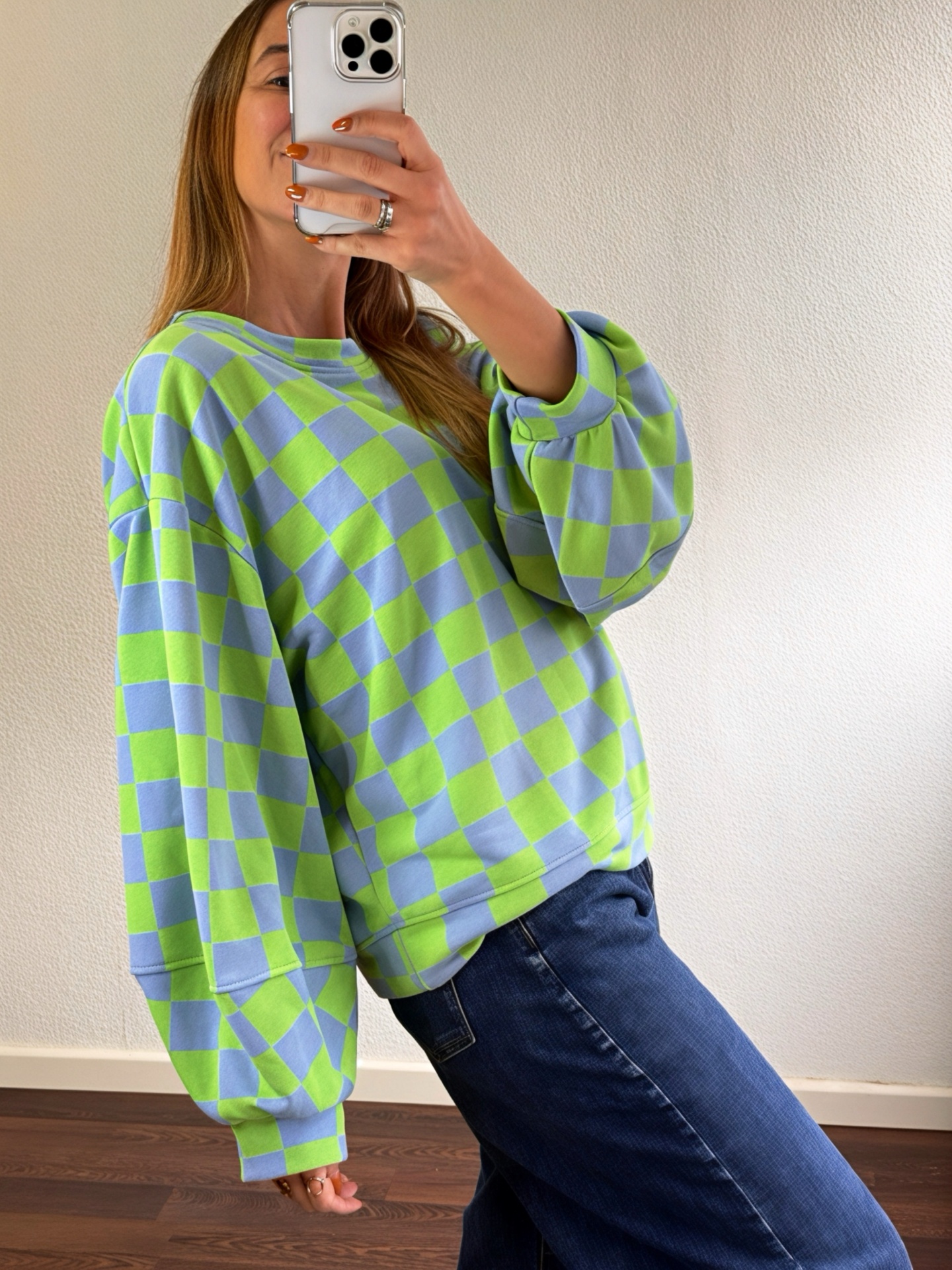 Pessoa a usar sweatshirt azul e verde claro com padrão de quadrados e calças de ganga azuis, tirada em interior com parede branca.