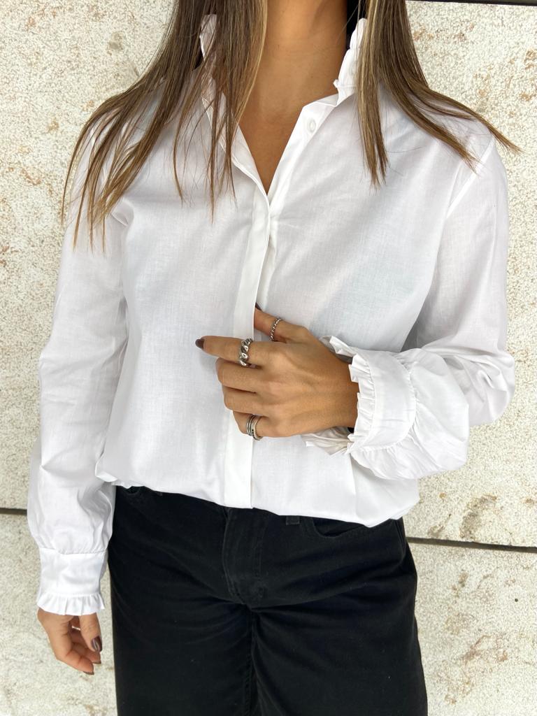 Blusa branca de manga comprida com punhos franzidos e gola detalhada, calças pretas, fundo claro texturado