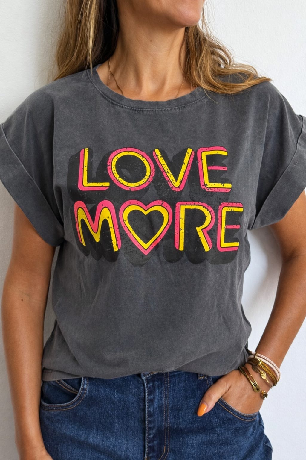 T-shirt cinza escuro com estampa LOVE MORE em amarelo e rosa e coração, uma pessoa veste jeans azul e pulseiras no pulso direito