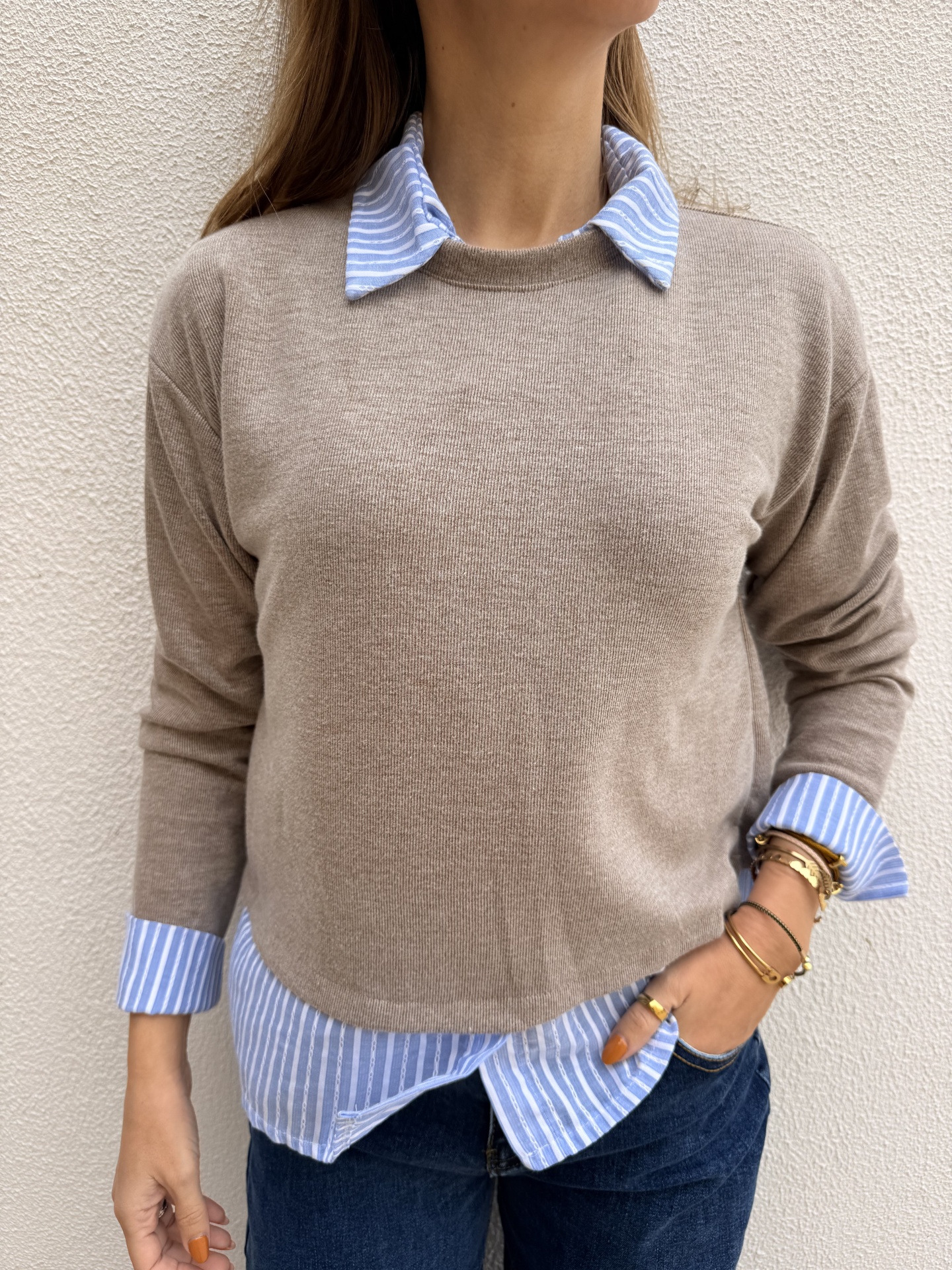 Pessoa com camisola bege sobre camisa azul riscada junto a parede clara