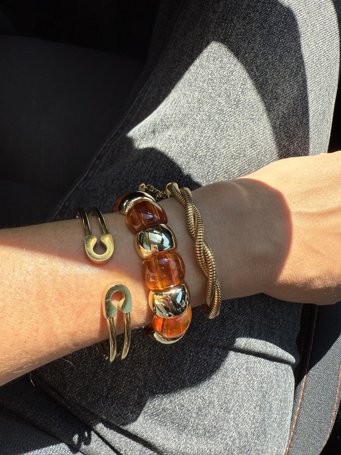 Três pulseiras douradas e castanhas no pulso com calças de ganga cinza ao fundo