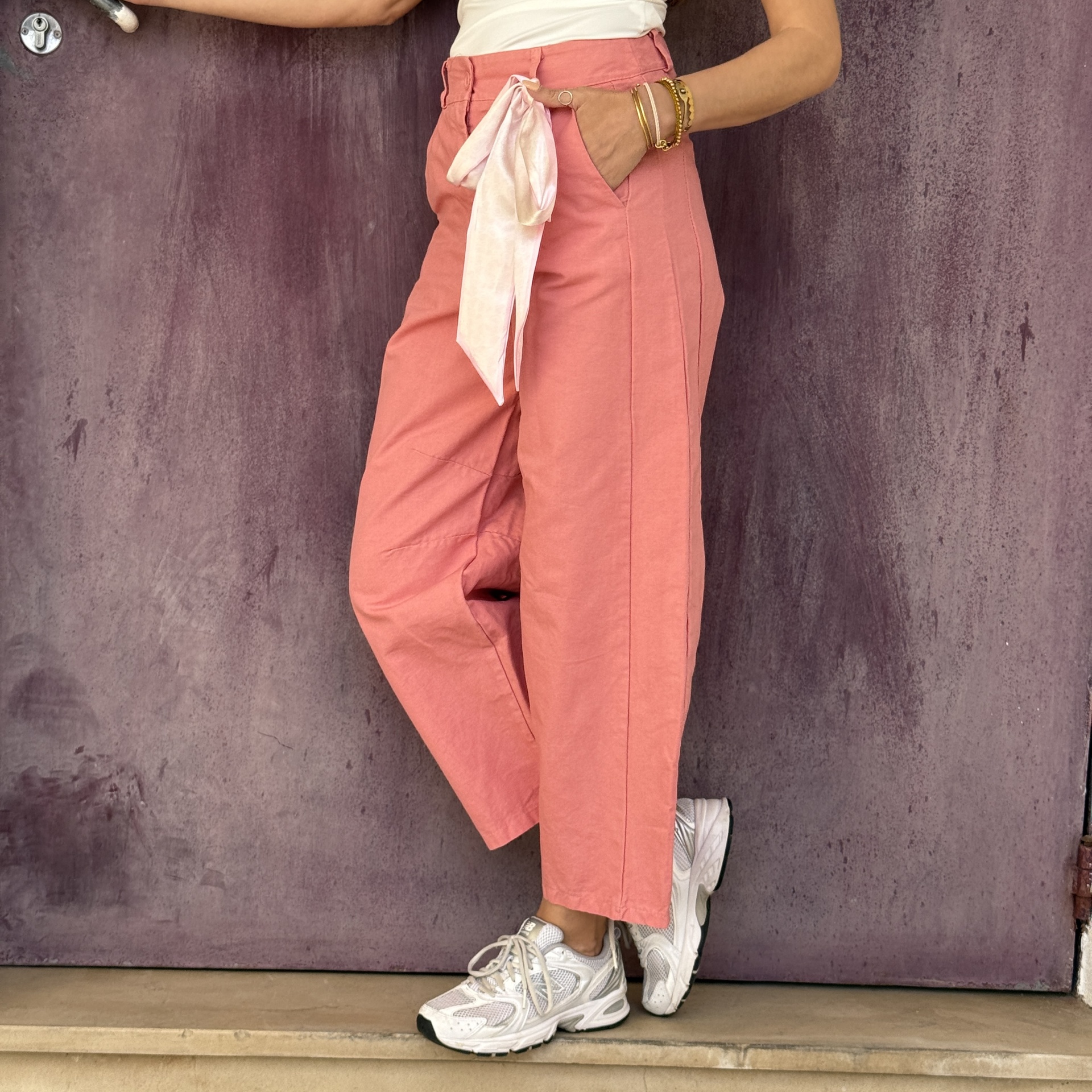 Calças rosa com laço rosa claro e ténis brancos em fundo de porta roxa