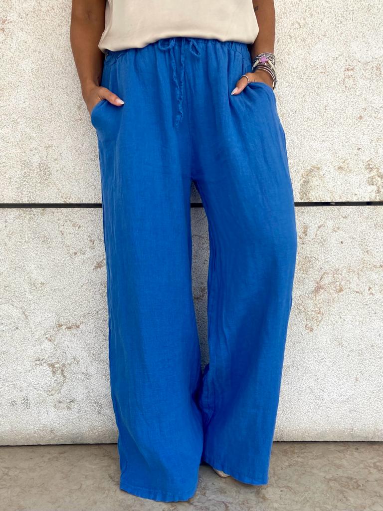 Calças largas azuis com bolsos e cós elástico contra parede de pedra clara