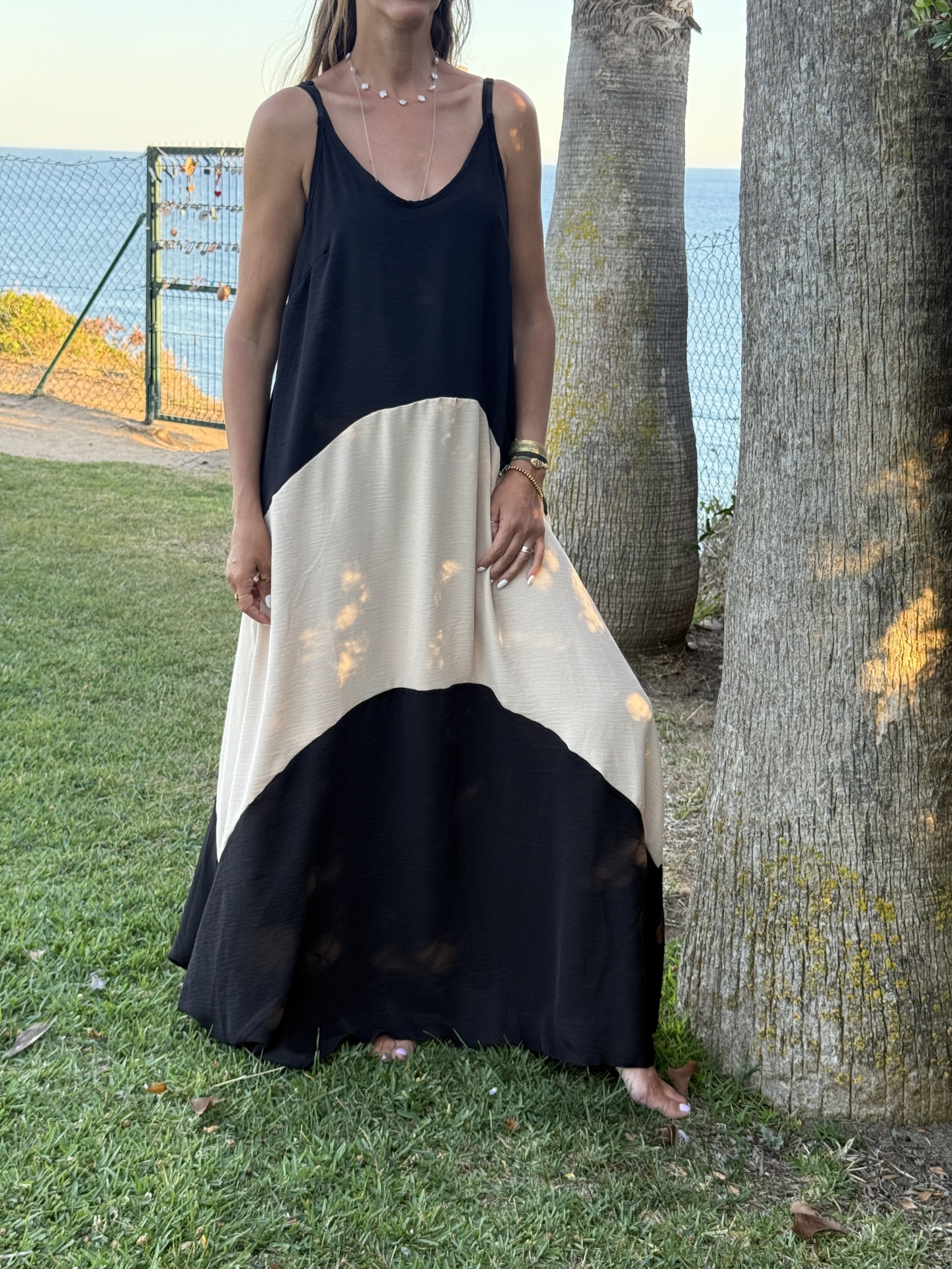 Mulher a usar vestido preto e bege em ambiente exterior com árvores e mar ao fundo