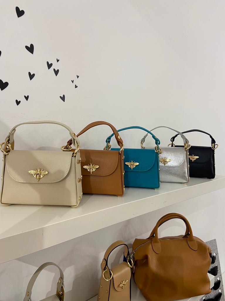 Malas de mão pequenas coloridas com abelhas douradas numa prateleira branca e malas maiores castanhas em baixo