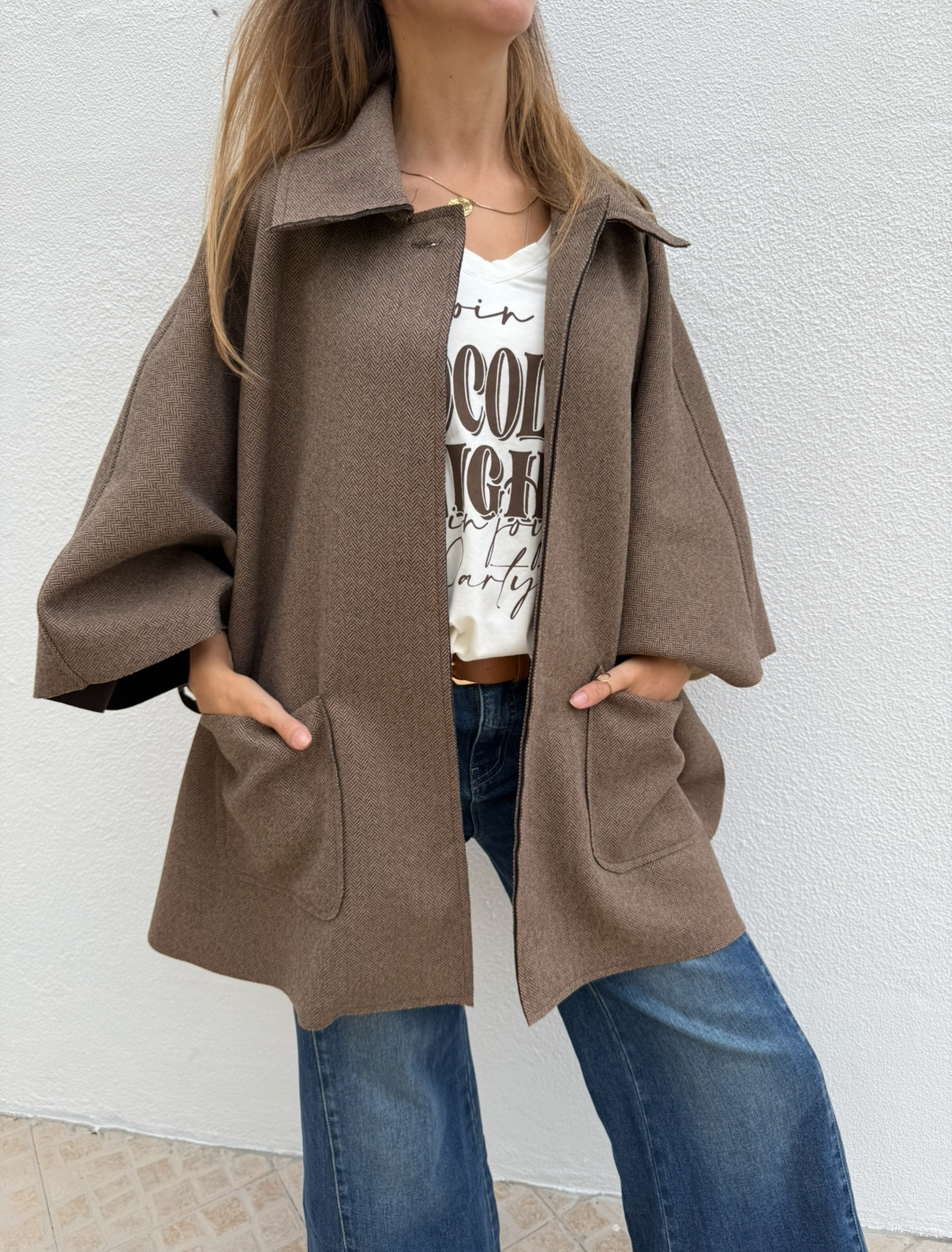 Casaco castanho com padrão espinha de peixe e bolsos, sobre t-shirt branca com texto e calças de ganga azuis.