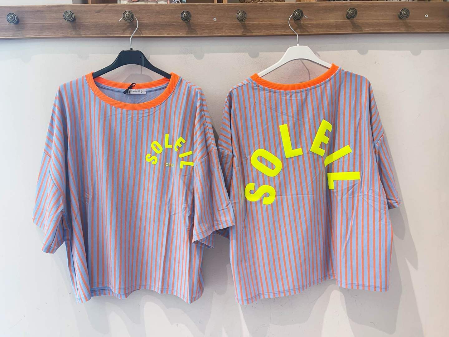 Duas t-shirts riscas laranja e azul com texto amarelo, penduradas numa parede