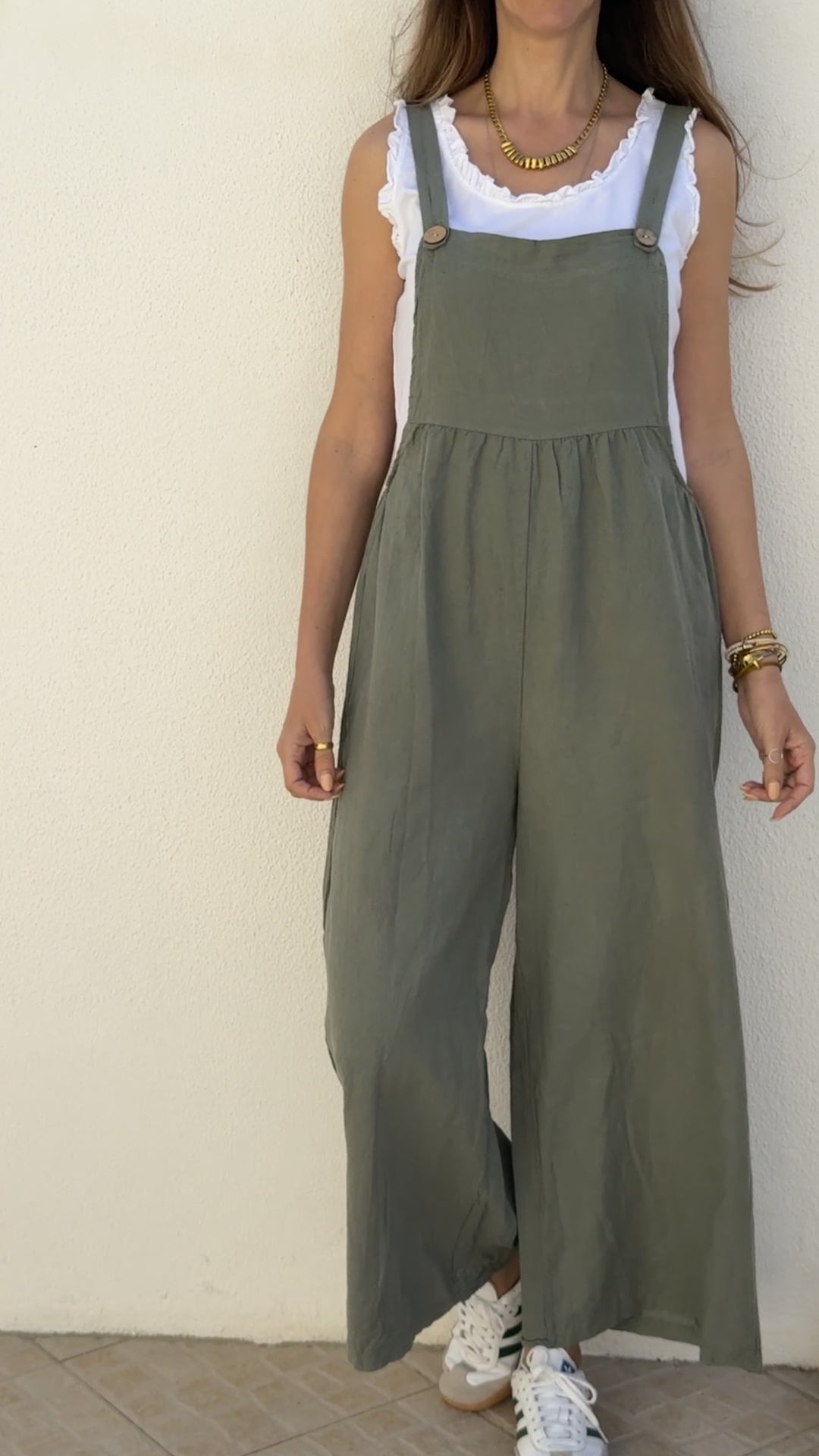 Macacão verde largo com alças e botões de madeira, sobre t-shirt branca de mangas com babados, com sapatilhas brancas de riscas verdes, parede branca ao fundo.