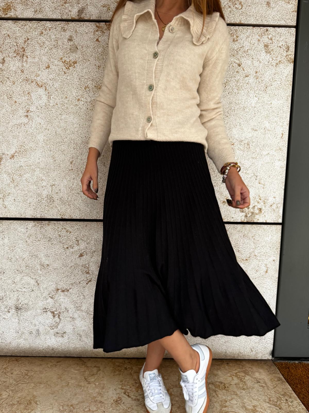 Cardigan bege malha, saia preta plissada midi, ténis brancos e castanhos, parede clara texturada