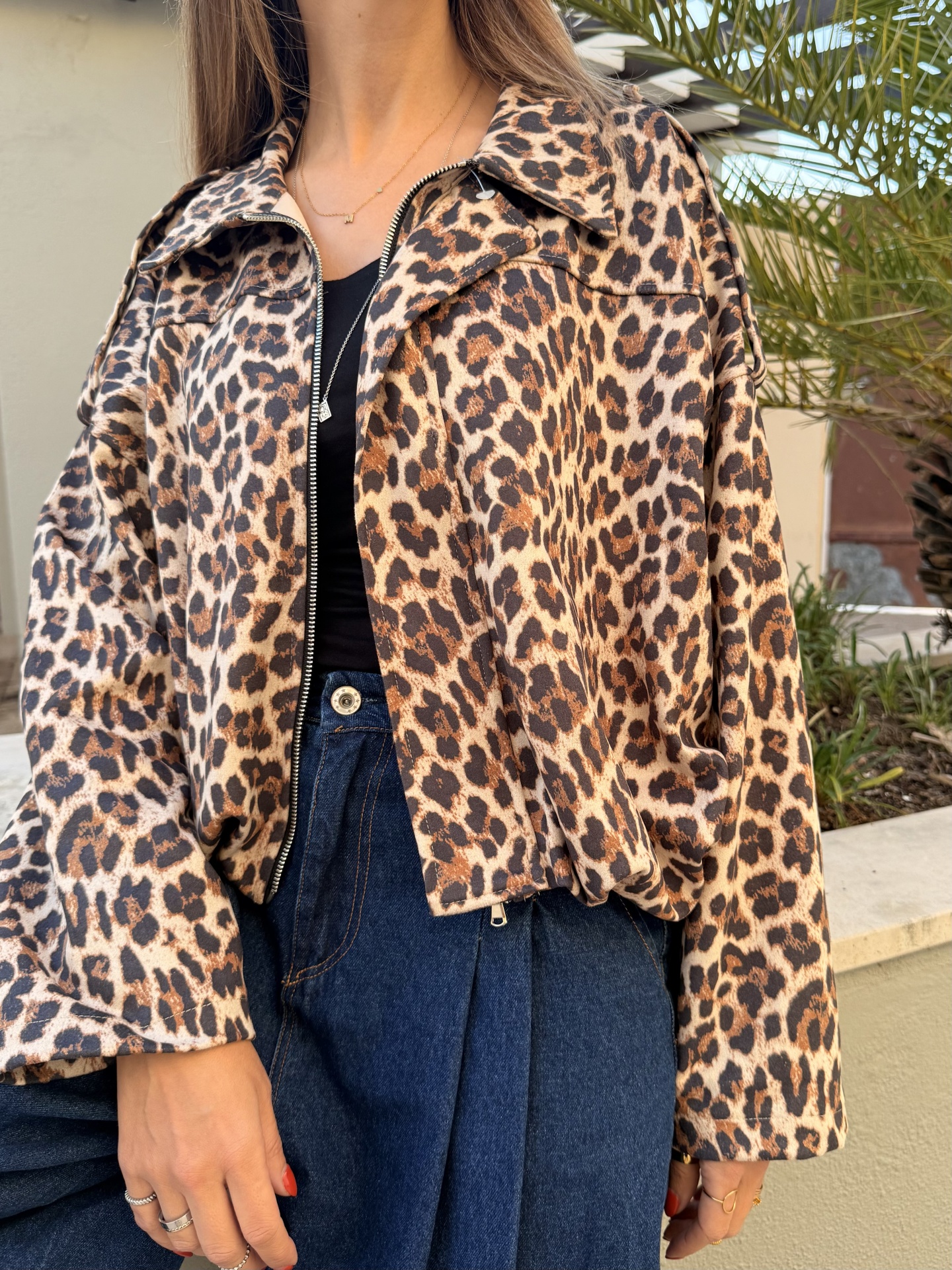 Casaco curto animal print castanho e preto com fecho de correr, calças de ganga azul e camisola preta num ambiente exterior com plantas.