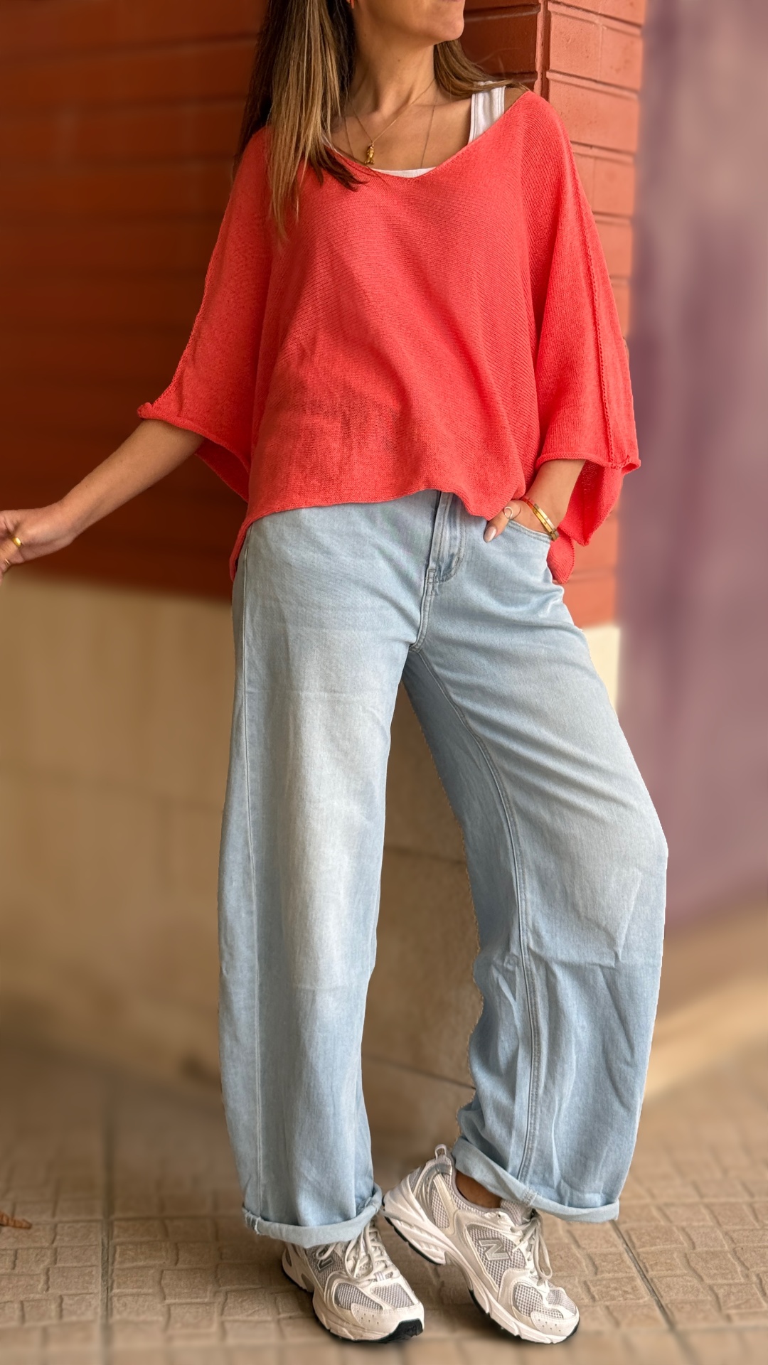 Pessoa com camisola larga coral, calças de ganga claras largas e ténis brancos em pavimento de azulejos